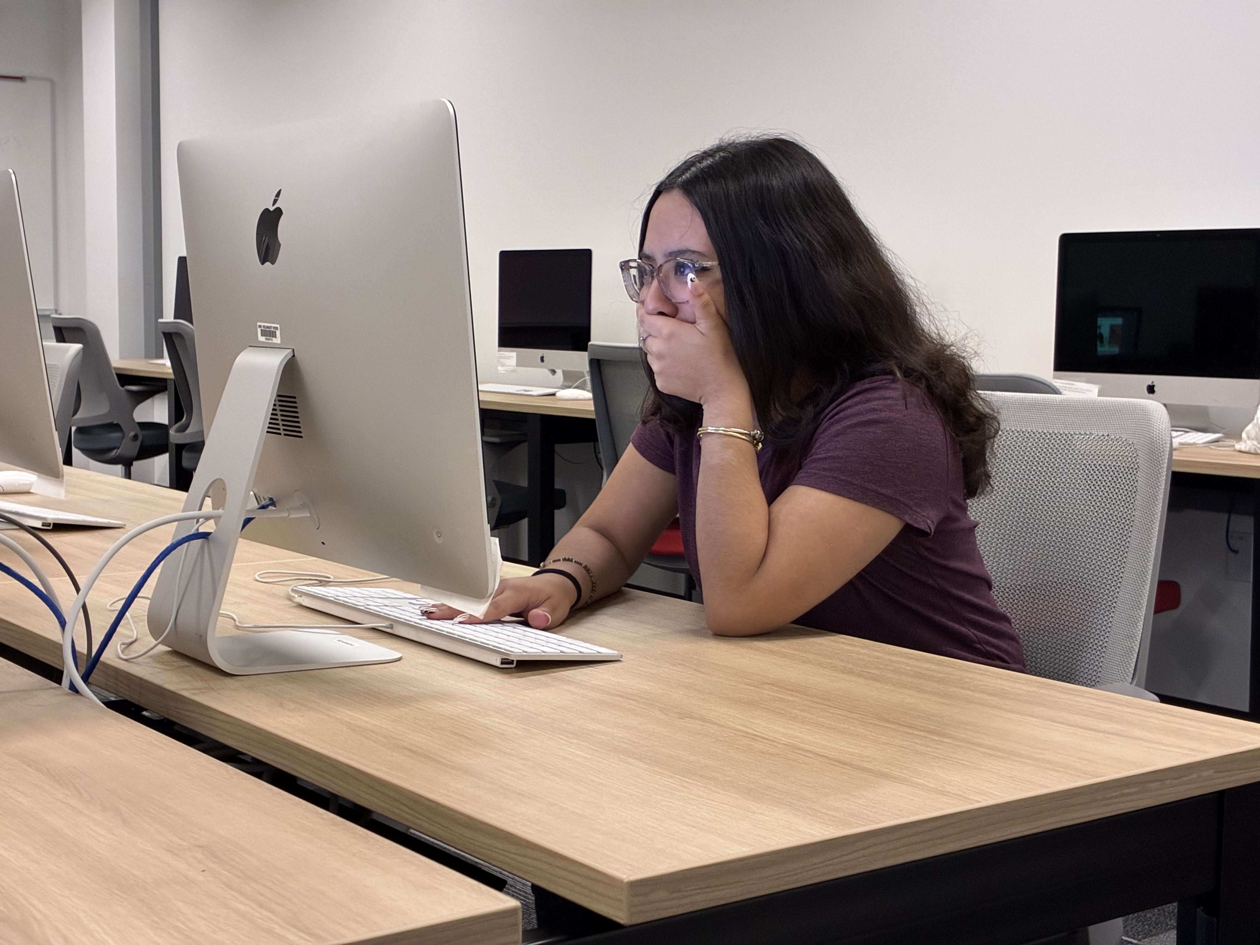 College student sits at a computer in a campus computer lab with her hand covering her mouth.